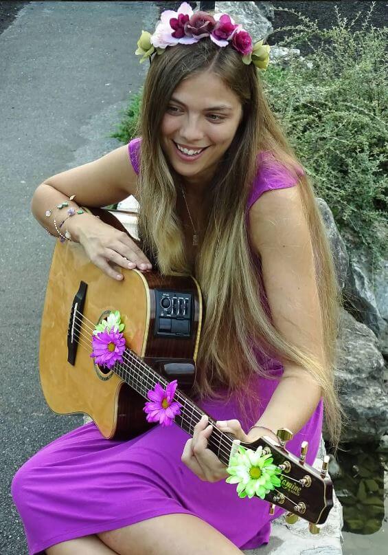 Amber Nadine in purple dress with flowers in hair playing guitar in seated position.