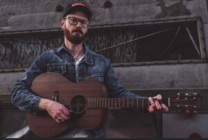 Braden McDannell holding guitar with wood beam in background.