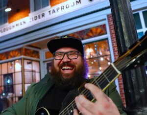 Shine Delphi holding guitar and laughing in front of brewery. 