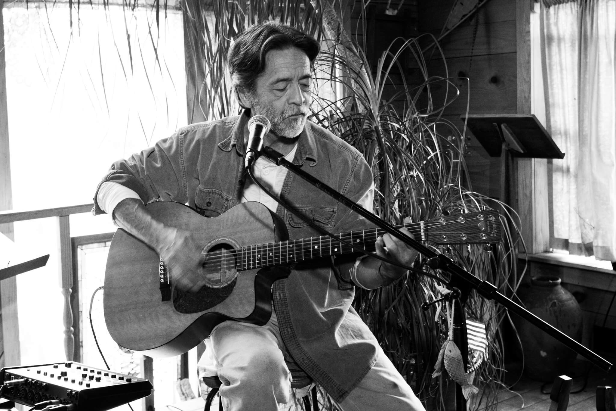 Black and white image of Stan Hoke playing guitar while seated and singing into microphone.