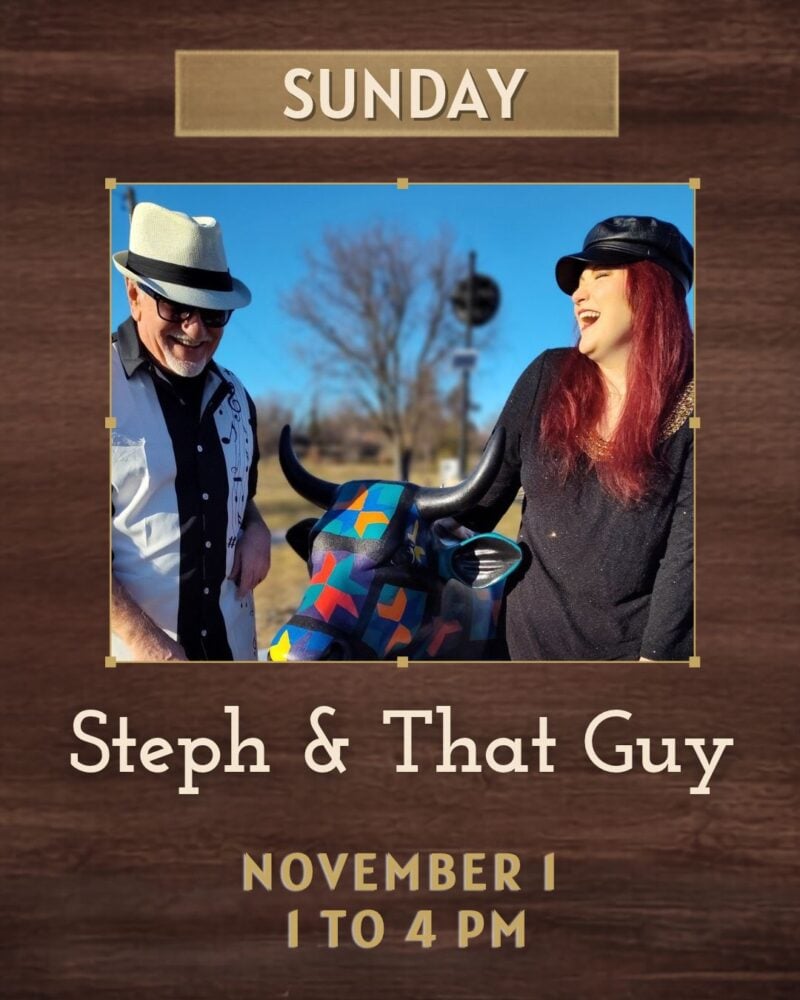 A man in a white fedora stands next to a woman in a black hat, black shirt and bright red hair outdoors. They are laughing together. A painted statue of a bull is between them.